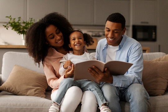 Story time. Loving African American father and mother reading interesting fairy tales book to his little daughter, girl enjoying time with parents, sitting on sofa at home
