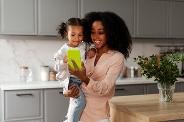 Happy African American mother and her adorable little daughter having video call via smartphone, spending time together at home, holding kid on her arms