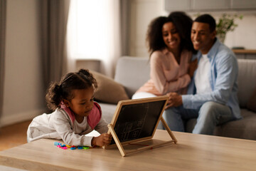 Pretty little girl playing with magnetic alphabet board while her happy African American parents looking at their daughter and smiling, sitting on sofa