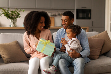 Daughter birthday surprise. Mom giving gift box greeting kid girl, child sitting on father's laps, parents and child celebrating holiday together