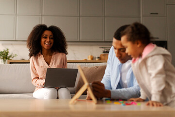 Black father engaging in early education with little daughter while mother working online on laptop and looking at husband and child. Family, parenting and professional life