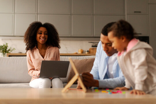 Black father engaging in early education with little daughter while mother working online on laptop and looking at husband and child. Family, parenting and professional life