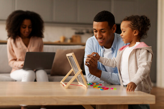 Family, parenting and professional life. Black father engaging in early education with little daughter while mother focusing on online work on laptop - Powered by Adobe