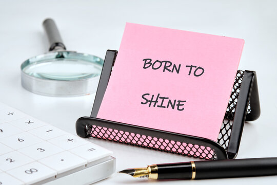 A bright pink note with the words born to shine sits neatly in a desk holder. Nearby, a pen, calculator, and magnifying glass create an inspiring workspace atmosphere, promoting creativity