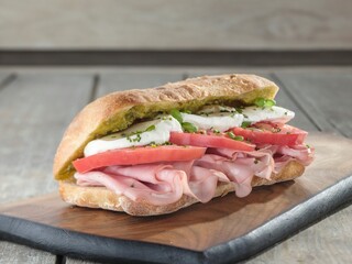 A delicious sandwich made with layers of creamy mozzarella, fresh tomatoes, and savory ham rests on a wooden board. The rustic setting enhances its appetite appeal