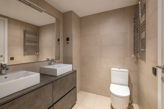 Modern bathroom with beige tiles, double sinks, a white toilet, and a large mirror. Clean and minimalist design with contemporary fixtures. - Powered by Adobe