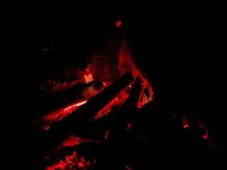 A striking close-up photograph captures the deep red heat and small flames of a smoldering campfire in complete darkness. The glowing embers and charred wood logs create a dramatic contrast against th