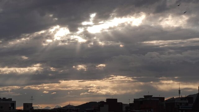 Dramatic sky with dark clouds and rays of sunlight breaking through. Urban skyline silhouette at dusk. Atmospheric and moody scene.
