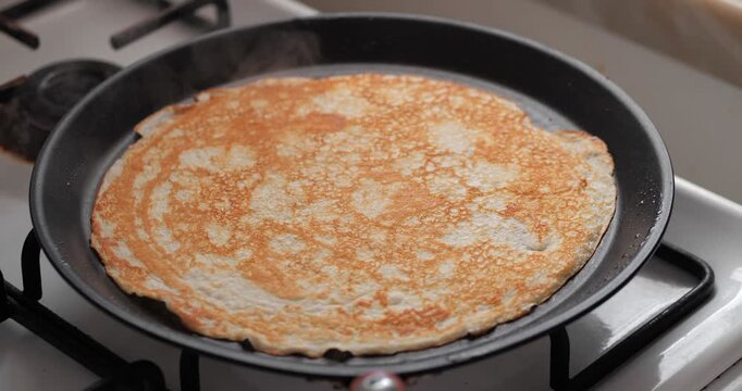 A golden-brown pancake cooking in a black frying pan on a gas stove. The pancake has a smooth texture and is evenly cooked.