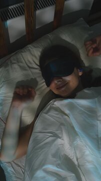 Vertical video. Slept and well rested woman lying in bed, taking off her sleep mask and earplugs, stretching arms, and waking up in a good mood at early morning.