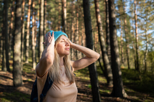 Relaxed inspired woman breathing deeply in sunlit forest with eyes closed hands on head. Grounding in wilderness, female stress relief in nature, slow conscious inhalation, mind and body unity