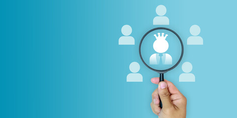 Hand holding magnifying glass focusing on a crowned figure among people icons on blue background,...