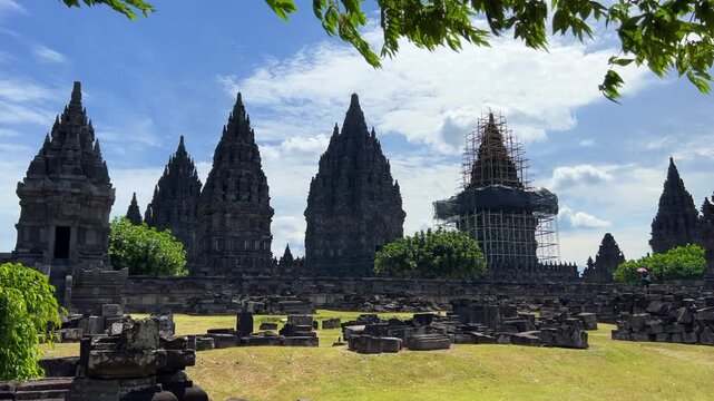 View of the ancient Prambanan Temple, a Hindu temple complex in Yogyakarta, Indonesia. The temple rises above the green foliage under a blue sky. The towers cast shadows on a sunny day. 4К