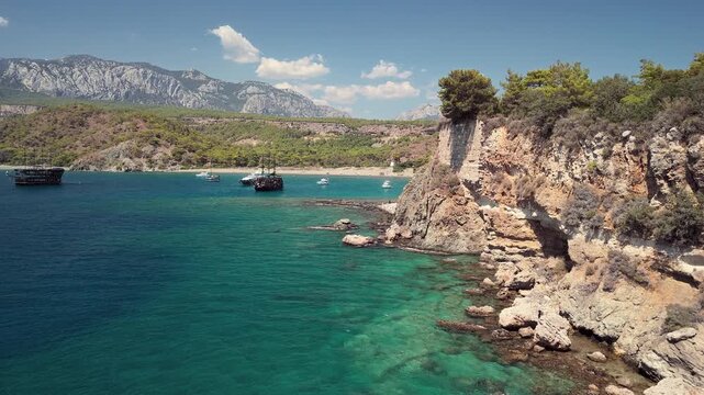 Aerial view of Phaselis Bay with sailing ships and Mount Olympos in the background. Clear blue water and scenic Mediterranean coastline near Antalya, Turkey. Captures scenic coastal beauty in Turkey.