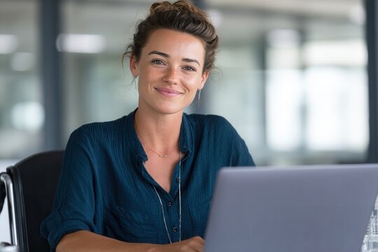 A cheerful woman sits at a desk with a laptop in a contemporary office setting, exuding confidence and professionalism.