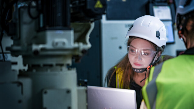 Young Asian female engineer in safety helmet using laptop to inspect industrial machinery in a modern factory, working closely with her team to ensure production quality.