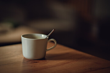 Empty white ceramic mug with metal spoon on wooden table in soft interior lighting.