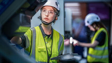 A female engineer in a hard hat and safety vest uses a tablet and stylus. She is focused, working in a high-tech smart factory or control room on a night shift.