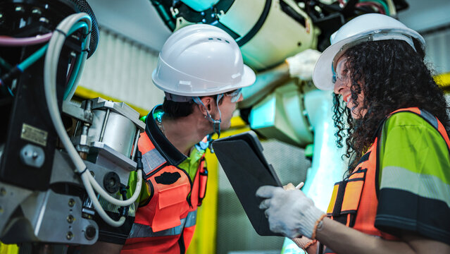 A male and female engineer in hard hats and safety vests collaborate on a high tech robotic arm. The woman holds a light while the man inspects a component in a smart factory.