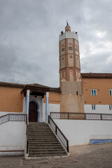 Fototapeta premium The Grand Mosque of Chefchaouen, Morocco, featuring its unique octagonal minaret under a cloudy sky. Stone steps lead to a decorative wooden entrance set into the terracotta-colored wall.