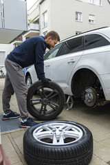 Man changing car tire using electric drill and jack on city sidewalk. DIY auto repair, maintenance, seasonal tire change, mechanical work, vehicle service, urban life.