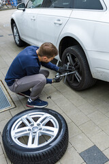 Man changing car tire using electric drill and jack on city sidewalk. DIY auto repair, maintenance, seasonal tire change, mechanical work, vehicle service, urban life.