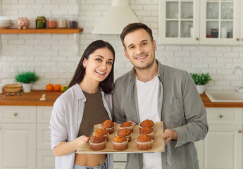 Happy young couple holding tray with yummy self baked tasty muffins out of oven, enjoy cooking in...