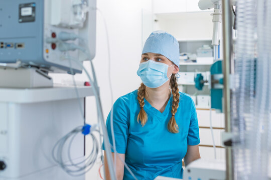 A healthcare worker in surgical scrubs is focused on a medical monitor in a bright hospital room during a procedure.