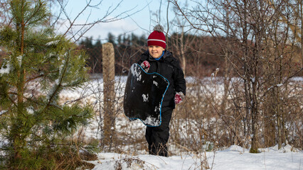 A little boy is having fun during the winter holidays and climbs a hill to slide down on his snowboard.