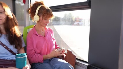Woman traveler sitting by the window in a city bus. She is plugging her smartphone into a usb port to charge it while listening to music - Powered by Adobe