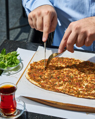 A lahmacun with a fork and knife next to a glass of tea and a plate of mint leaves