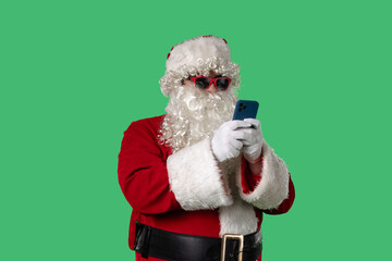 Santa Claus with a white curly beard, wearing a red suit and sunglasses, texting on a smartphone on a green background. Perfect for Christmas-themed compositions, advertising, and holiday layouts.