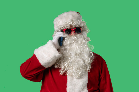 Santa Claus with a white curly beard, wearing a red suit and sunglasses, talking on a smartphone against a green background. Perfect for Christmas-themed compositions, advertising, and holiday layout - Powered by Adobe