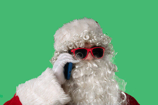 Santa Claus with a white curly beard, wearing a red suit and sunglasses, talking on a smartphone against a green background. Perfect for Christmas-themed compositions, advertising, and holiday layout