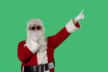 Santa Claus with a white curly beard, wearing a red suit and sunglasses, points his finger on a green background. Perfect for Christmas-themed compositions, advertising, and holiday layouts.