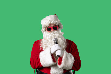 Santa Claus with a white curly beard, wearing a red suit and sunglasses, sings songs into a microphone against a green background. Ideal for Christmas themed compositions and holiday layouts.