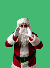 Santa Claus with a white curly beard, wearing a red suit and sunglasses, is shouting with his hands cupped around his mouth against a green background. Ideal for Christmas themed compositions.