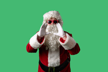 Santa Claus with a white curly beard, wearing a red suit and sunglasses, is shouting with his hands cupped around his mouth against a green background. Ideal for Christmas themed compositions.