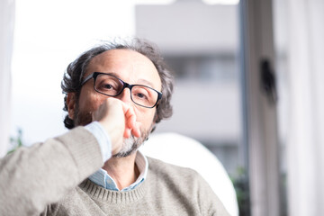 Portrait of a thoughtful middle-aged man with glasses sitting indoors by a window. Casual sweater, soft light, introspective mood. Concept of reflection, thinking or modern lifestyle.
