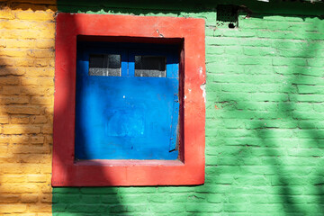 A window in the middle of a brick wall with various vibrant colors, shadows cast by trees not shown in the image, La Boca, Buenos Aires