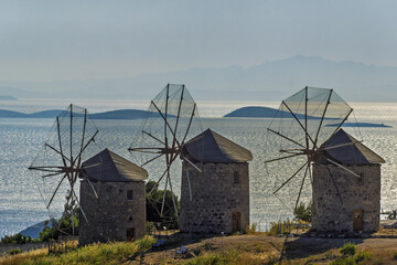 Three traditional stone windmills with conical roofs and wooden sails standing on grassy terrain near the sea, aligned in a row facing the water, with small islands in Patmos, Greece