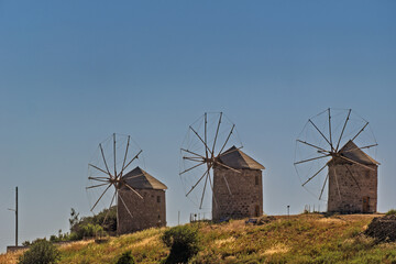 Three traditional stone windmills with conical roofs and wooden sails standing on grassy terrain near the sea, aligned in a row facing the water, with small islands in Patmos, Greece