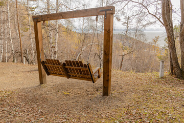 Wooden bench swing hangs from a sturdy frame in a quiet autumn park overlook distant hills and a hazy valley and a river or lake beyond the trees.