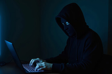 A hacker in a black mask works on a laptop at night. A masked hacker hacks websites in the dark, wearing a hood. Anonymous hacker in a mask breaking into computer systems in a dark room.