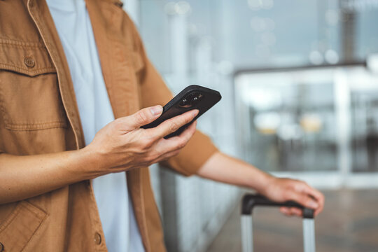 Close up of asian man hand holding smartphone while waiting with luggage at airport terminal, modern travel lifestyle concept, technology connection during journey, and vacation preparation.