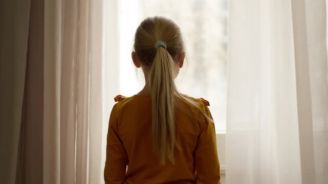 Back view Caucasian little girl child kid daughter looking out window at street outside dreaming contemplate waiting family parents leaving room walking going away at cozy home baby offspring lonely