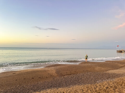 Man fishing on beach at sunset, catching fish rod into calm sea, outdoor adventure, relaxing vacation, fisherman silhouette, fishing hobby concept. Fisherman for pike, perch, carp.