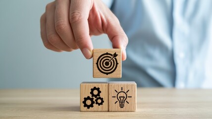 Hand stacks wooden blocks with icons for goals, gears, and ideas, symbolizing strategy and planning.