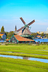 A traditional windmill and historic wooden houses on the waterfront of a lake or wide river, with green fields in the foreground.