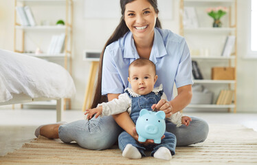 A smiling mother sits on the floor with her baby in a bright nursery, holding a blue piggybank to teach early savings and parental financial skills. Encouraging early saving habits at home.